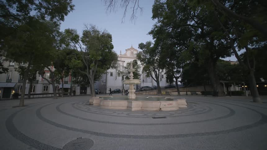 A square with a fountain in the center surrounded by a concrete floor with some trees and a white building in the background