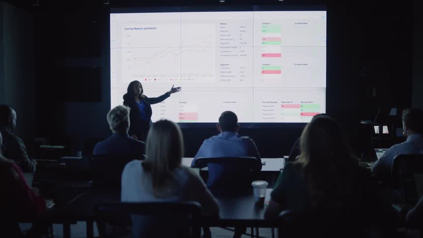 A woman does a presentation in a large room full of people sitting at desks using a large wall screen