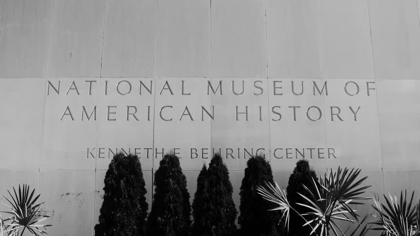 The entrance of the national museum of american history featuring the buildings name engraved on the wall is framed by tall shrubs and plants