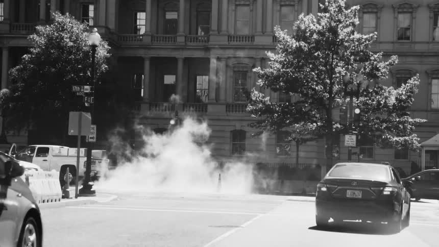 Black and white street scene with a car driving on a street with steam rising