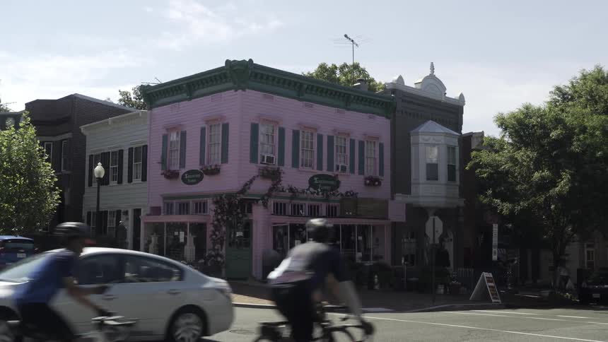 A group of cyclists riding past a charming twostory corner building with pink walls green shutters and decorative plants on its faade located on a quiet street with trees and neighboring buildings