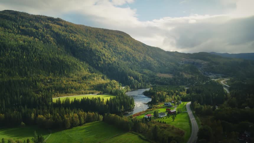 A winding river flows through a lush valley surrounded by forested hills with scattered houses and a road leading through the green landscape under a partly cloudy sky