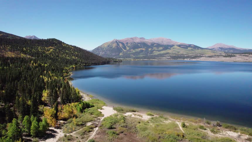 Panoramic view of a mountainous landscape with a large body of water in the foreground surrounded by dense forests and mountains in the background under a clear blue sky