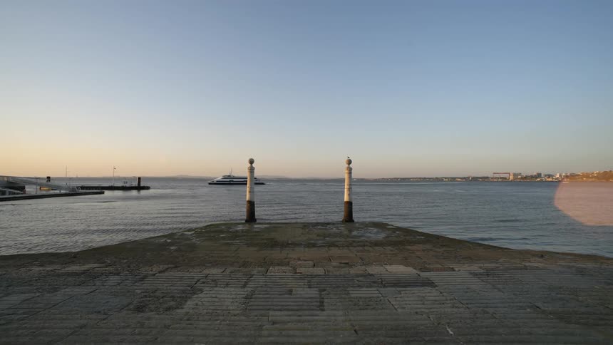 Waterfront scene with two posts at the end of a paved promenade and a ferry sailing behind
