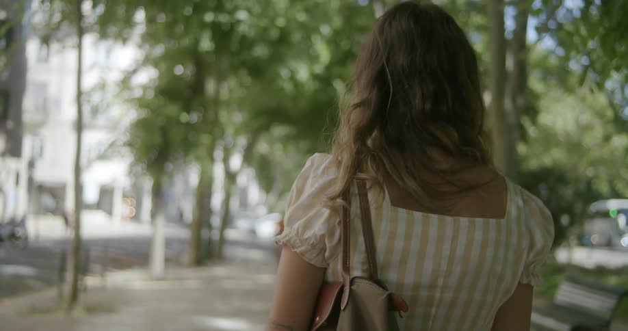 A person with long hair is walking down a treelined street during the day