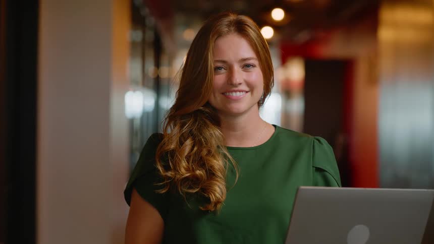 Woman holding a laptop and giving a smile while standing in a hallway