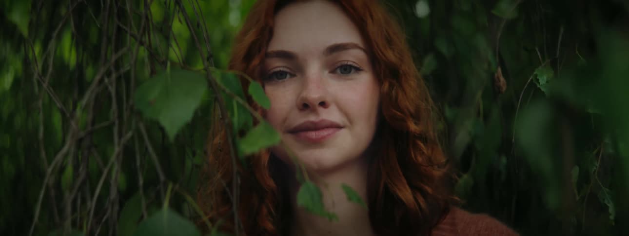 The face of a redhaired woman with a subtle smile and she is behind some green leaves