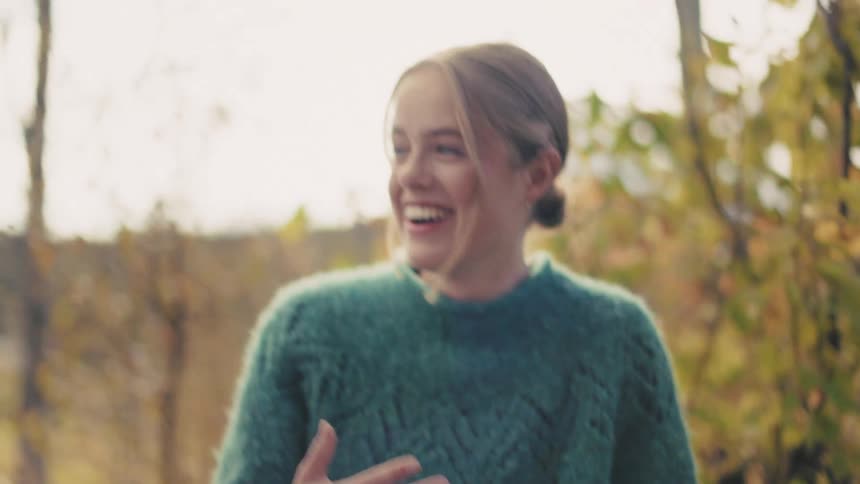 A blonde woman sitting outside is happily smiling and showing her hand with a golden ring
