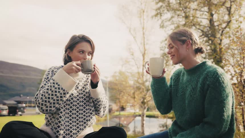 Two women sitting outdoors each holding a mug toasting and drinking