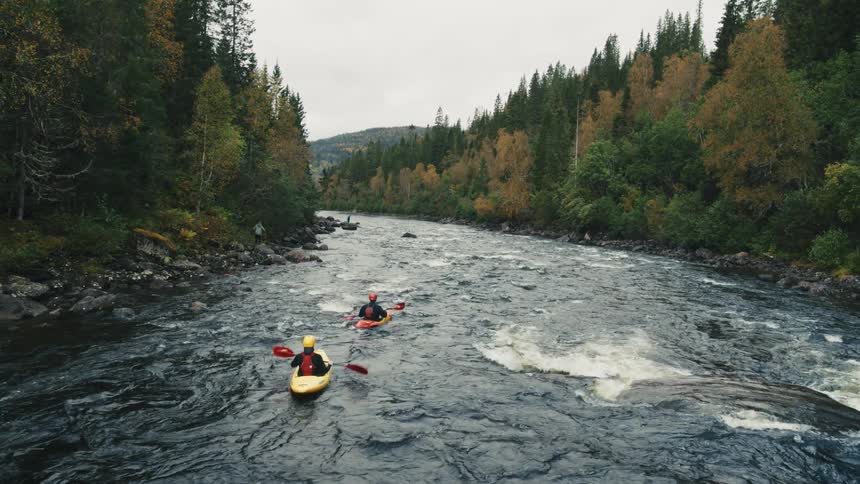 Two individuals kayaking on a river with rapid currents surrounded by dense forest with autumncolored foliage