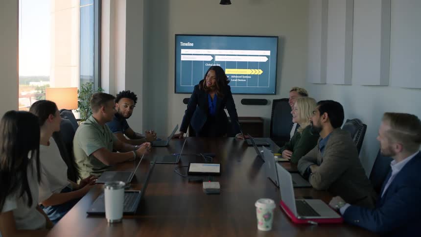A manager in a corporate meeting walks to a desk where a group is seated and listens to one of the team members talk