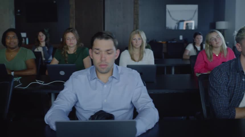 Group of individuals seated at a long table in a conference room with laptops open
