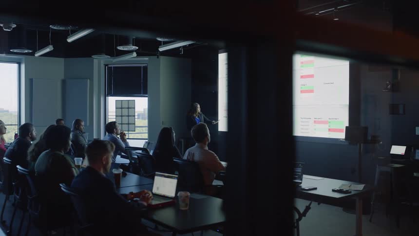 Person giving a presentation in a modern office meeting room with attendees seated and looking towards a large screen