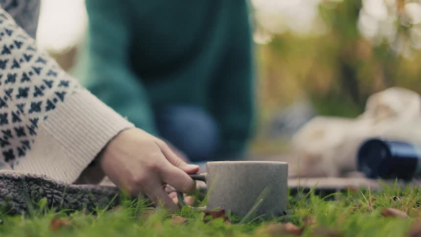 Woman picking up a mug on the grass and in the background another person sitting reading a book