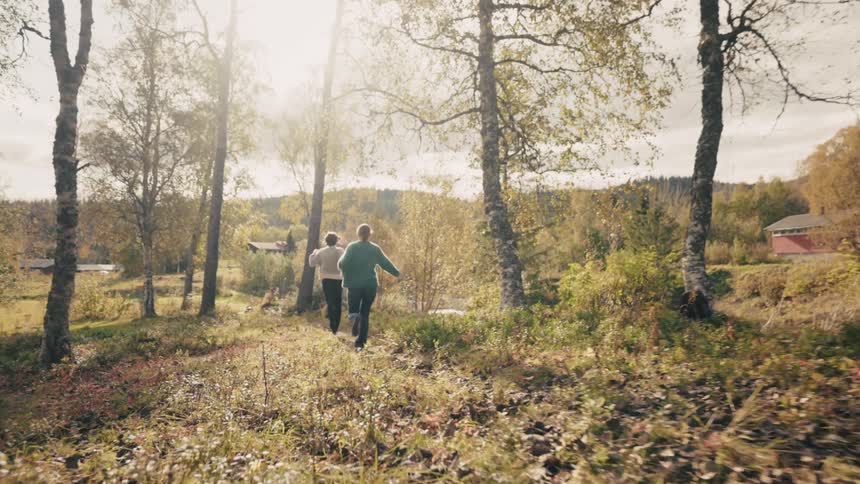 Two friends running through a sunlit forest with trees displaying autumn colors
