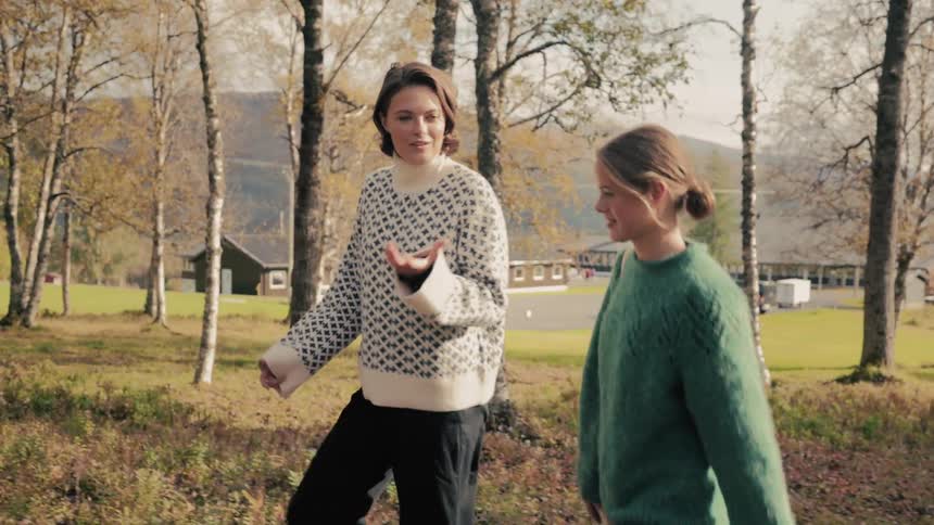 Two women one wearing a white patterned sweater and the other in a green sweater are walking up a hill on a fall day talking as they pass through a park with trees and colorful autumn scenery