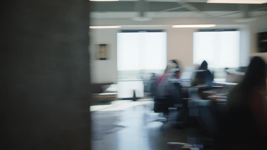 A woman pushing a cart with a box of paper on top and in front there is a large office table with several people working on laptops