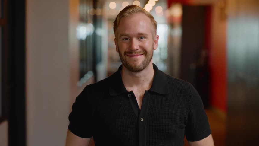 A man with a beard smiles warmly standing in a hallway