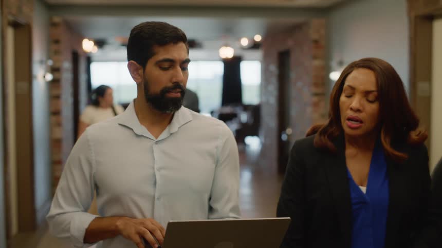 A group of coworkers exits an elevator walking down a hallway together talking and looking at a laptop