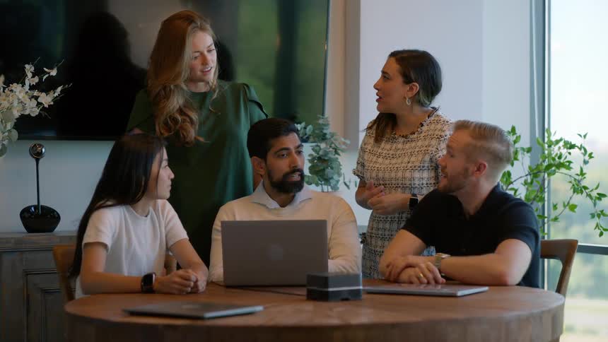 Group of five people at an office desk and facing a laptop talking to each other