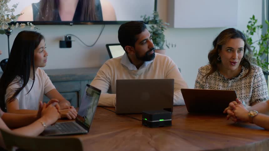 A group of some people gathered around a round wooden table with laptops and they are paying attention to a woman who is talking