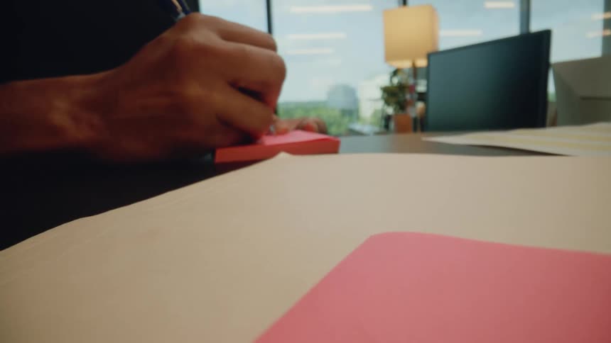 A hand writes on a pink sticky note while sitting at a desk