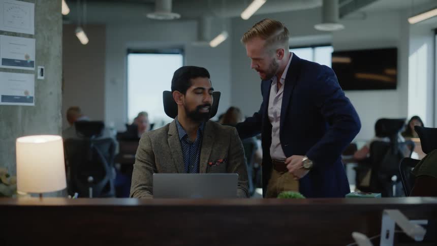 A man gets up from his desk and walks to another man to collaborate over a laptop