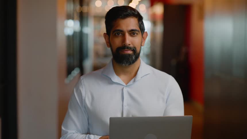 Bearded man in button up shirt holding a laptop in a hallway