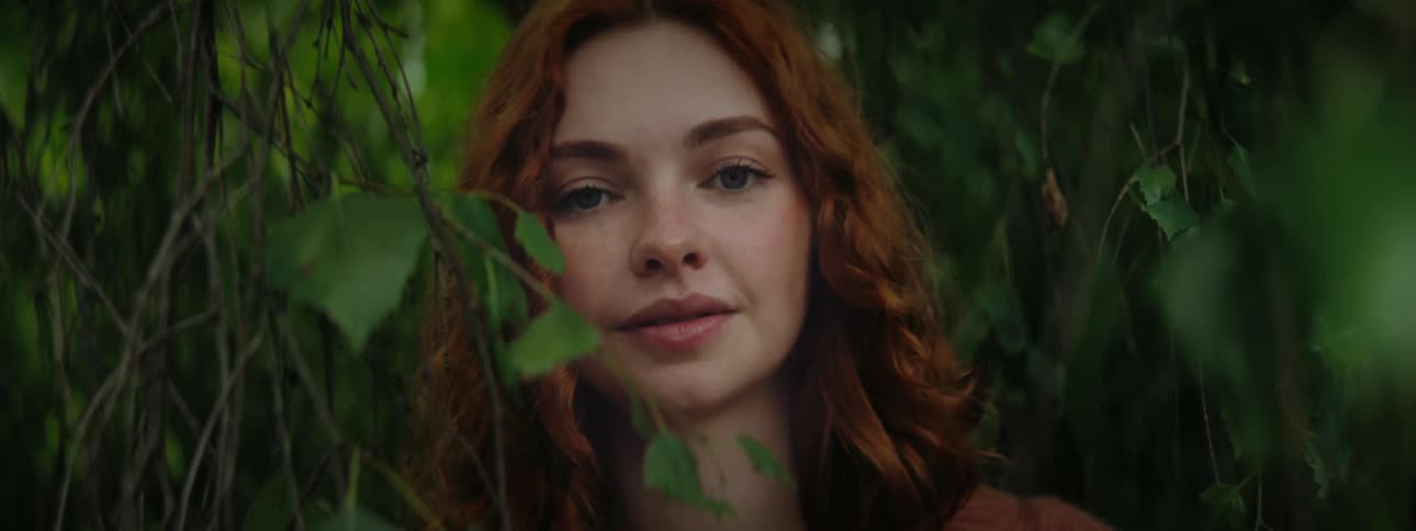 A woman with long red hair peeks through a thicket of green leaves