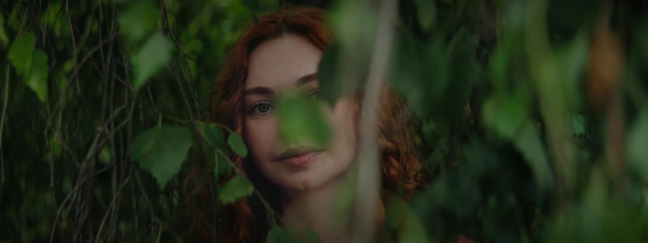 A young woman with red hair partially concealed by green foliage