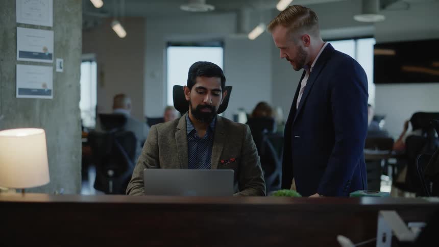 A man walks over to another man who is seated in a large office setting