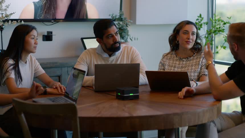 People sitting at a table in a modern workspace talking in an inperson and virtual meeting