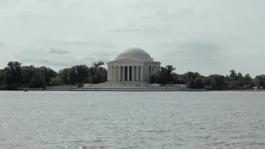 The jefferson memorial stands by the water with gentle waves as people walk around the monument surrounded by trees