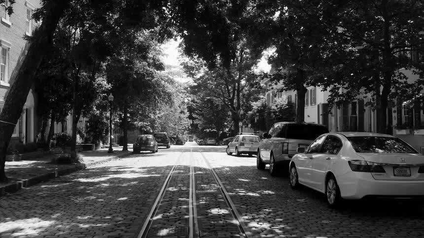 A cobblestone street with a rail in the middle and some cars parked on the sides in black and white tones