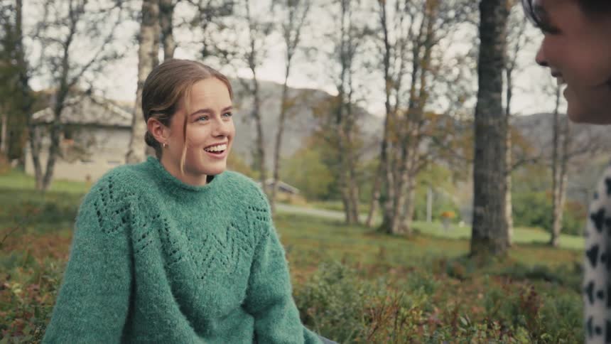 Two women sitting on grass with trees around smiling talking