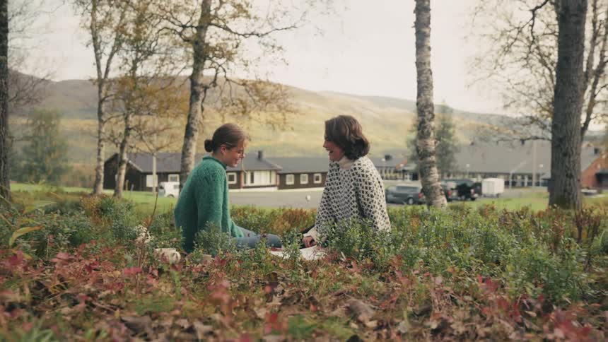 Two women laughing while sitting on the grass with mountains in the background