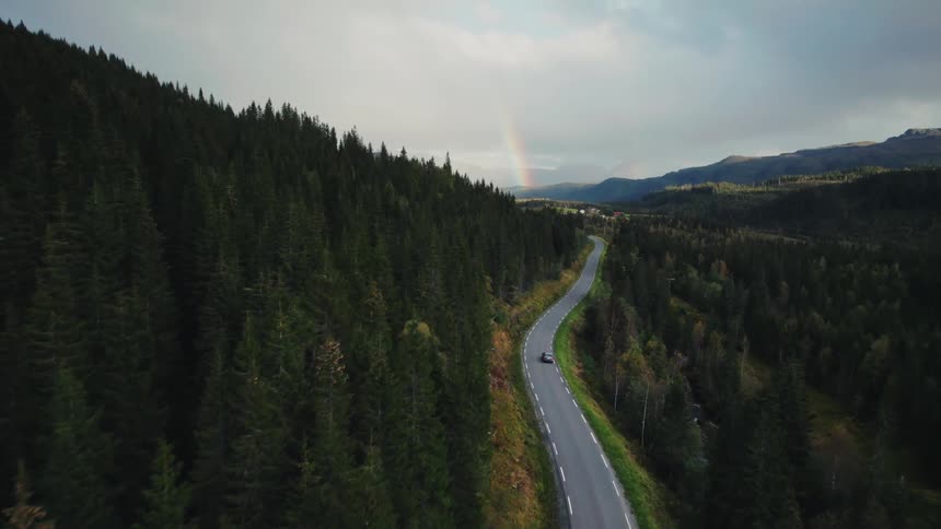 Aerial view of a car driving on a winding road cutting through a dense forest with a rainbow in the distance