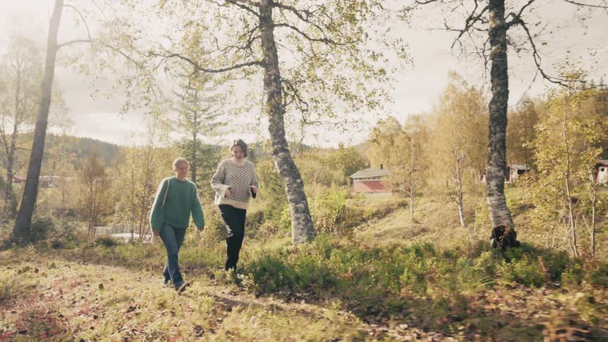 Two women walking through a rural landscape with trees and small structures in the background