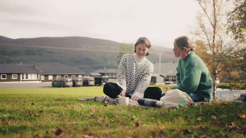 Two friends sitting on a cloth in the grass of a field laughing and talking with parked cars and mountains in the background