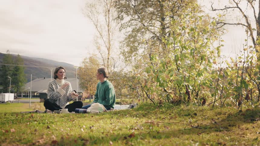 Two women sitting on the grass in a park setting with trees and a mountain in the background