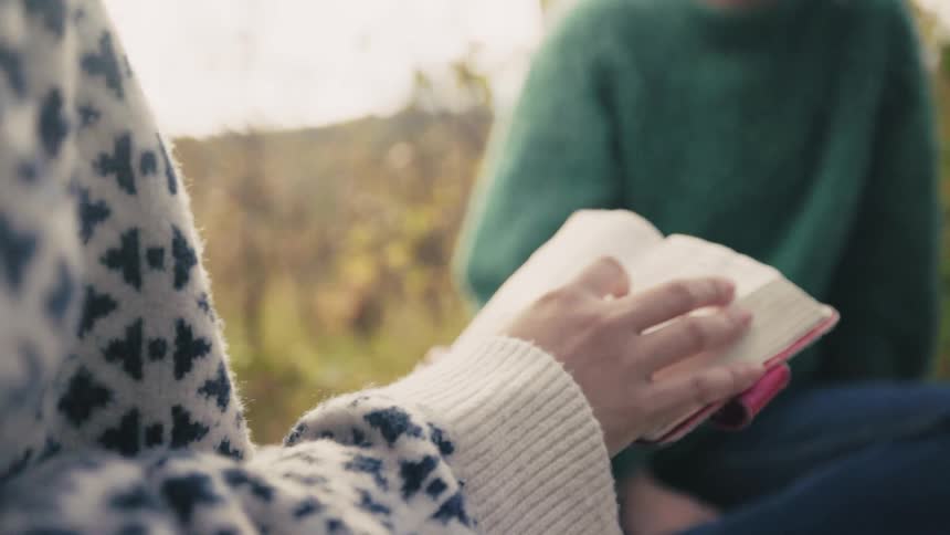 Two friends wearing thick sweaters reading a book outdoors