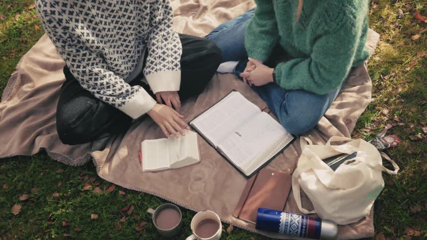 Two individuals seated on a blanket outdoors with an open book and cups of hot beverages