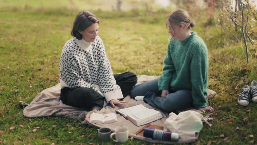 Two friends sitting on a cloth in the grass with books talking to each other