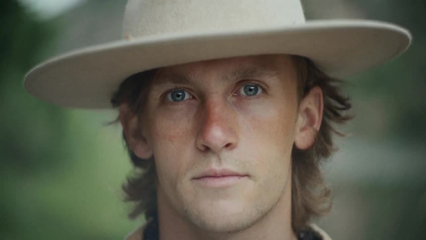 A man wearing a flat brimmed hat stands on a grassy plain in nature