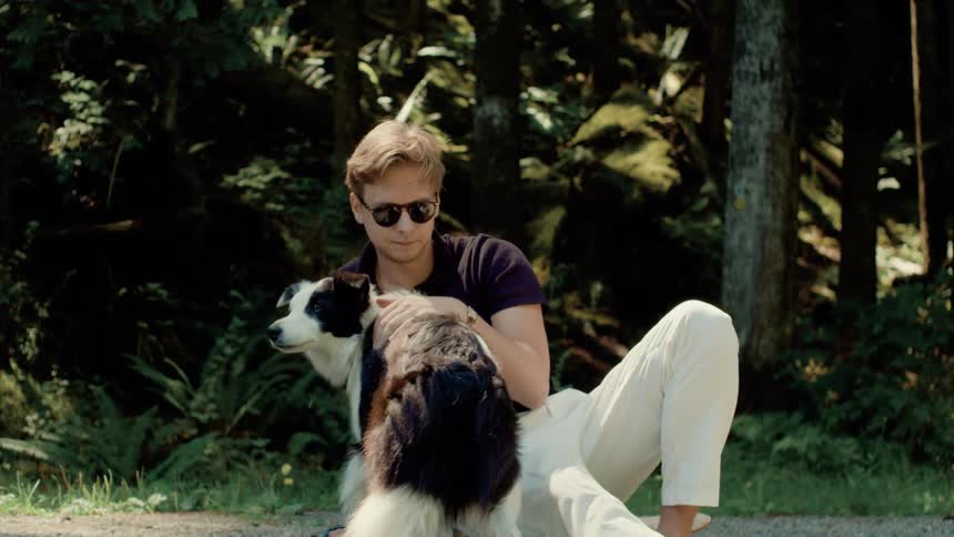 A man in sunglasses reclines outdoors while petting the back of a dog