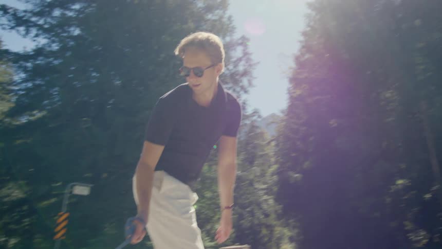 A man in a black tshirt and white pants walks his dog in a park while riding a skateboard on a sunny day