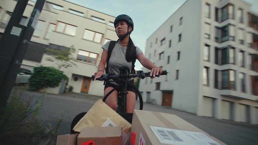 Female cyclist pedals a cargo bicycle containing packages for delivery past apartment buildings