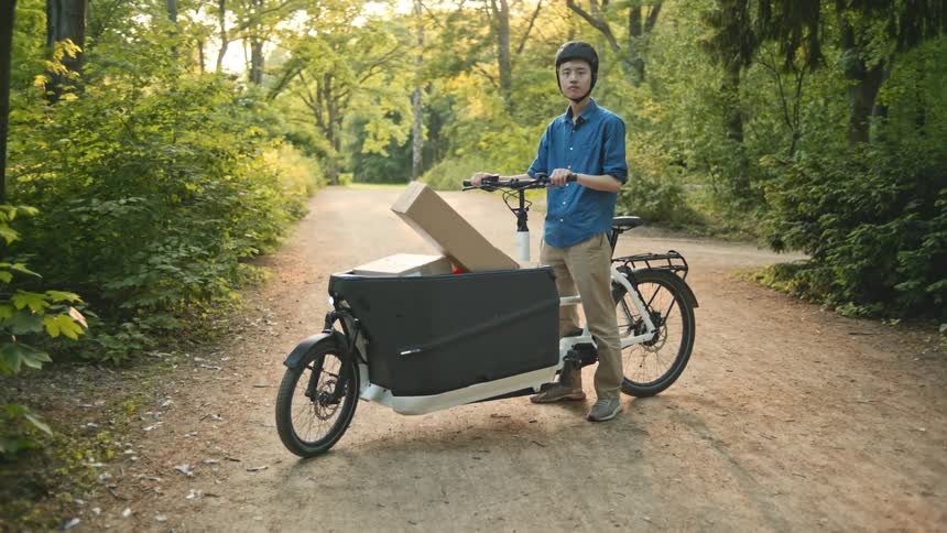 Teenager on a bicycle carrying boxes in the middle of a dirt road surrounded by trees