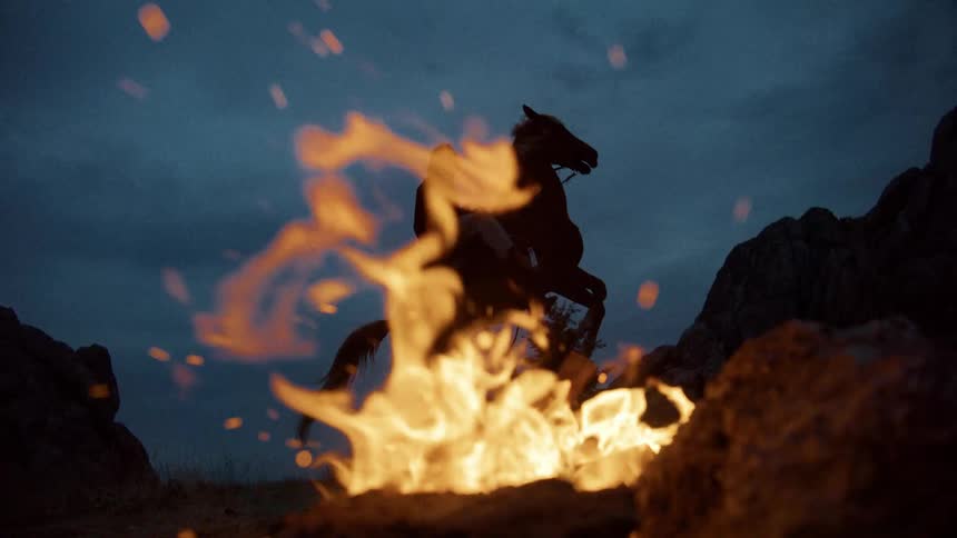 A man on horseback silhouetted against a dramatic sunset with a bonfire burning in the foreground
