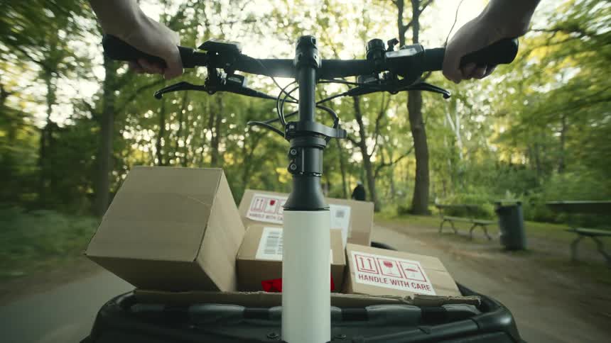 A person riding a bike with a cargo carrier full of boxes labeled handle with care on a sunny day in a park POV